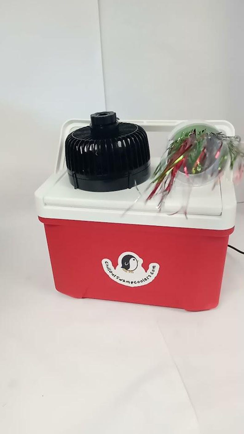 May include: Red and white cooler with a black fan on top. A clear container with colorful streamers sits on the cooler. The cooler has a white handle and a sticker with a penguin logo and the text "chilloutswampcoolers.com".
