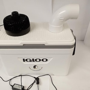 May include: A white Igloo cooler with a black fan and a white PVC pipe elbow on top. The cooler has a gray stripe and the Igloo logo. A power adapter and a black cord are connected to the cooler.