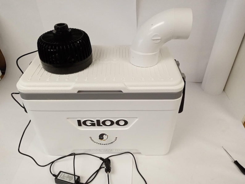 May include: A white Igloo cooler with a black fan and a white PVC pipe elbow on top. The cooler has a gray stripe and the Igloo logo. A power adapter and a black cord are connected to the cooler.