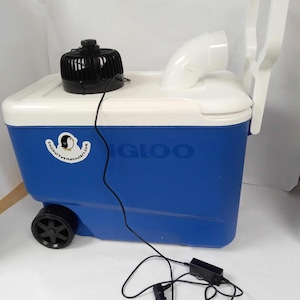 May include: A blue and white Igloo cooler with a black fan, white handle, and a white curved vent. The cooler has a black wheel and a power cord with a car adapter. The cooler has the Igloo logo on the side.
