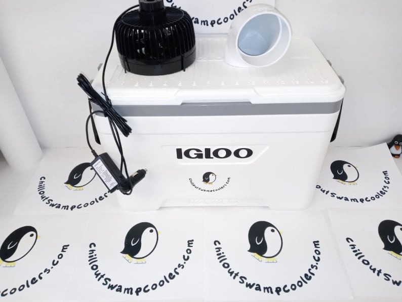 May include: A white Igloo cooler with a black fan and a white air vent on top. The cooler has a gray trim and the text "IGLOO" on the front. The image also shows a power adapter and several logos with a penguin.