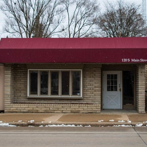 May include: Exterior view of a small brick building with a burgundy awning. The building has a large window and a white door. The text "120 S Main Street" is printed on the awning. Snow is falling.