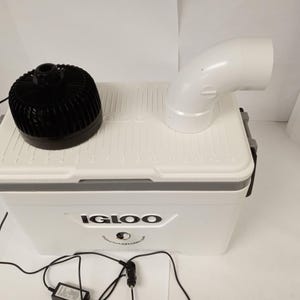 May include: A white Igloo cooler with a black fan and a white curved pipe attached to the top. The cooler has the Igloo logo on the front and is connected to a power source.