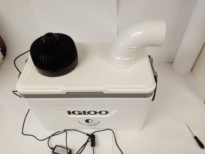 May include: A white Igloo cooler with a black fan and a white curved pipe attached to the top. The cooler has the Igloo logo on the front and is connected to a power source.