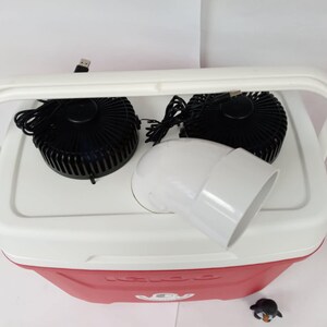May include: A white cooler with a red lid and a black handle. Two black fans are attached to the top of the cooler, and a white plastic pipe is connected to the fans. The cooler has the word "KOOL" printed on the lid.
