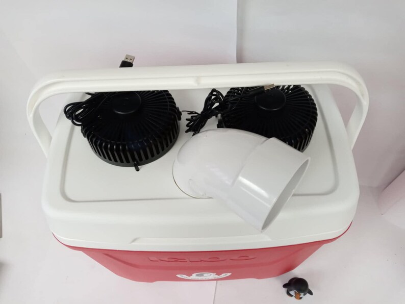 May include: A white cooler with a red lid and a black handle. Two black fans are attached to the top of the cooler, and a white plastic pipe is connected to the fans. The cooler has the word "KOOL" printed on the lid.