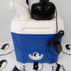 Peut inclure: Glaci&egrave;re Igloo bleue et blanche avec un ventilateur noir et un raccord de ventilation blanc. La glaci&egrave;re a un cordon d'alimentation noir et un autocollant avec un logo de pingouin et le texte "Chillout Swamp Coolers.com". Une petite figurine de pingouin est devant.