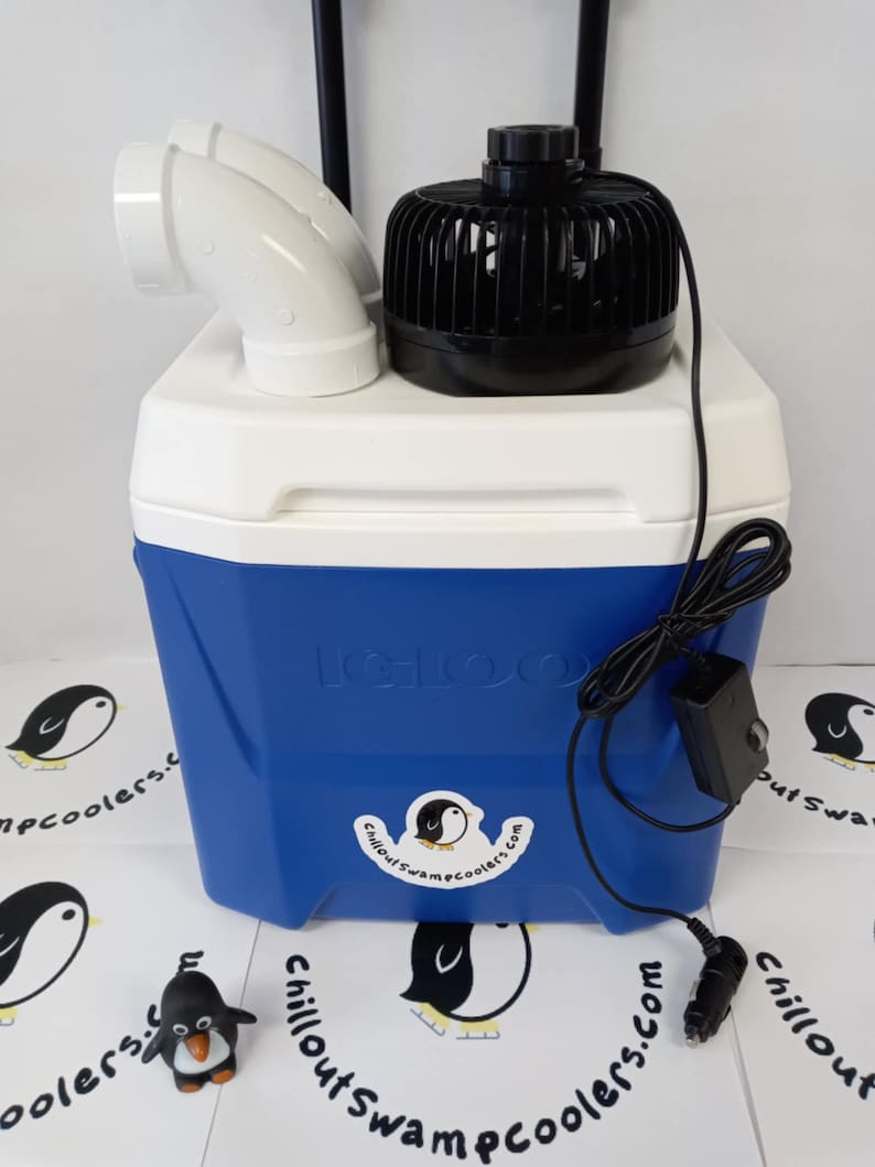 Peut inclure: Glaci&egrave;re Igloo bleue et blanche avec un ventilateur noir et un raccord de ventilation blanc. La glaci&egrave;re a un cordon d'alimentation noir et un autocollant avec un logo de pingouin et le texte "Chillout Swamp Coolers.com". Une petite figurine de pingouin est devant.