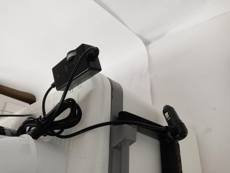 Puede incluir: Un adaptador de corriente negro con una perilla y un cable conectado a una nevera blanca con un asa negra.