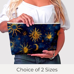 May include: A dark blue cosmetic bag with a celestial design featuring gold suns, moons, and stars. The bag has a gold zipper and is held by a person. The text "Choice of 2 Sizes" is at the bottom.