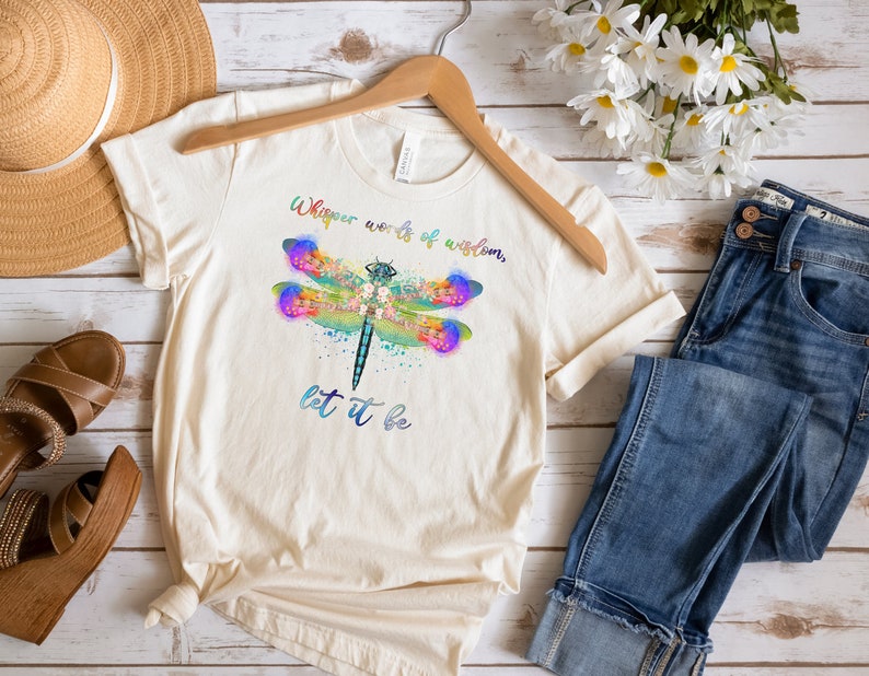 Puede incluir: Una camiseta de color crema con un dise&ntilde;o de lib&eacute;lula acuarela y el texto "Whisper words of wisdom, let it be".