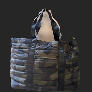 Peut inclure: Un grand sac fourre-tout matelassé avec un motif camouflage dans les tons de vert, marron et gris. Le sac a des poignées noires et une bandoulière noire. Le sac est de forme rectangulaire et possède une fermeture éclair.