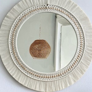 May include: A white macrame mirror with a fringe border and a round mirror inside. The mirror is reflecting a light fixture with a woven shade.