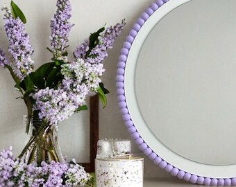 Op de afbeelding: Een ronde witte spiegel met een lavendelkleurige parelrand, die op een witte commode met twee lades staat. Twee boeketten lavendel staan aan weerszijden van de spiegel.