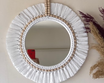 May include: White macrame mirror with a wooden bead border and a tassel hanging from the top. The mirror is round and has a white fringe around the edge.