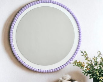Op de afbeelding: Een ronde spiegel met een witte lijst en een lavendelkleurige parelrand. De spiegel hangt aan een witte muur boven een houten tafel.