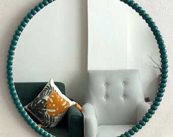 May include: A round mirror with a teal beaded frame. The mirror is reflecting a living room with a gray armchair and a green sofa with a pillow.
