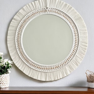 May include: A round mirror with a white macrame frame and a beaded border. The mirror is hanging on a white wall above a wooden shelf.