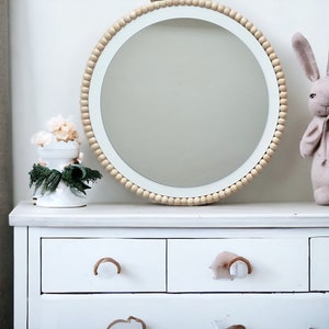 May include: White dresser with three drawers and wooden knobs. A round mirror with a wooden bead frame sits on top of the dresser. A small bouquet of flowers and a stuffed bunny are also on the dresser.
