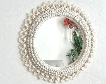May include: A round mirror with a white macrame frame. The frame is made of knotted rope with fringe details. The mirror is hanging on a white wall.