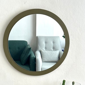 May include: A round mirror with a dark green frame hangs on a white wall. The mirror reflects two chairs, one green and one white, and a white wall. A glass terrarium with sand and various cacti sits on a wooden table.