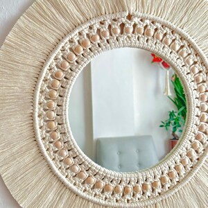 May include: A round mirror with a macrame frame and wooden beads. The frame is cream colored and has a fringe around the edge. The mirror is reflecting a room with a couch and plants.
