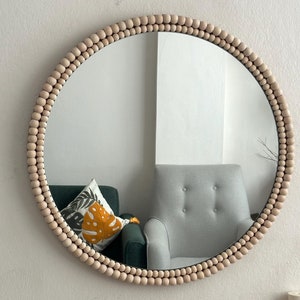 Pode incluir: Um espelho redondo com uma moldura de contas de madeira. As contas são de cor bege claro e estão dispostas em um padrão circular ao redor do espelho. O espelho reflete uma sala com uma cadeira verde e uma poltrona cinza.