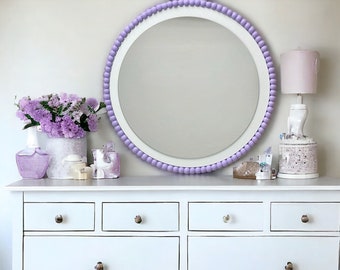 Op de afbeelding: Een witte commode met zes laden en zilveren knoppen. De commode is versierd met een paarse kralenspiegel, een vaas met paarse bloemen en een lamp met een roze lampenkap.