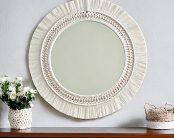 May include: A round mirror with a white macrame frame and a beaded border. The mirror is hanging on a white wall above a wooden shelf.