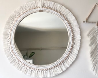 May include: A white round mirror with a fringe border and a beaded trim. The mirror is hanging on a white wall. A white macrame feather wall hanging is also hanging on the wall.