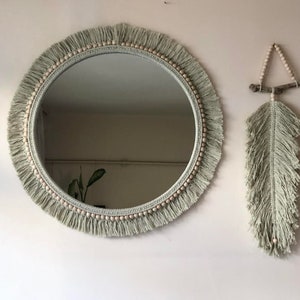 May include: A round mirror with a light green fringe and wooden beads around the edge. A light green macrame feather wall hanging is to the right of the mirror.