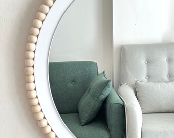Op de afbeelding: Een ronde witte spiegel met een houten kralenrand. De spiegel reflecteert twee fauteuils, een groene en een lichtgrijze. De spiegel is aan een witte muur bevestigd.