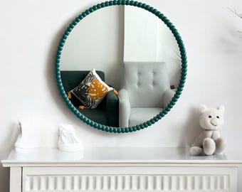 May include: A round mirror with a teal beaded frame hangs on a white wall above a white dresser. The mirror reflects a green armchair with a patterned pillow and a white armchair.