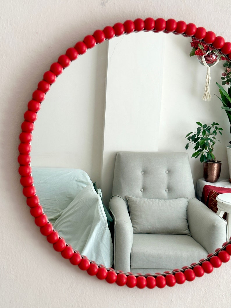May include: Round mirror with a vibrant red beaded frame. The mirror reflects a light gray armchair and other home decor items. The frame is made of small, evenly spaced, round beads.