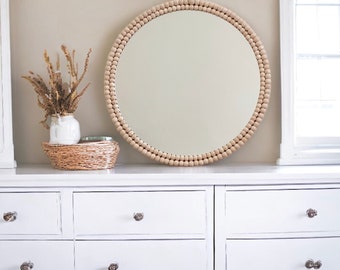 Op de afbeelding: Een witte commode met drie lades en een ronde spiegel met een houten kralenlijst. De spiegel hangt aan de muur boven de commode. Er staat een vaas met gedroogde bloemen op de commode.