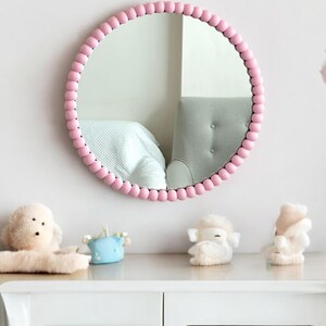 May include: A round mirror with a pink beaded frame hangs on a white wall. Below the mirror, a white dresser displays plush toys. The mirror reflects a bed and a gray chair. Decorative items hang on the wall.
