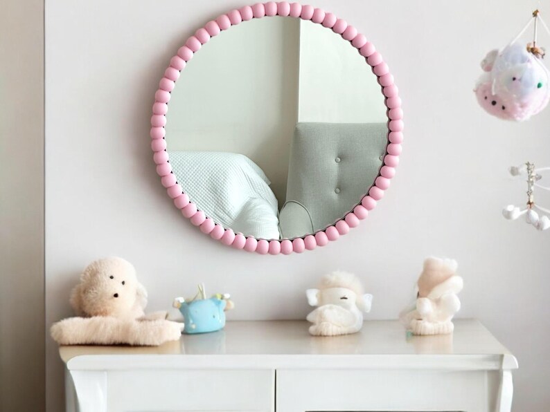 May include: A round mirror with a pink beaded frame hangs on a white wall. Below the mirror, a white dresser displays plush toys. The mirror reflects a bed and a gray chair. Decorative items hang on the wall.