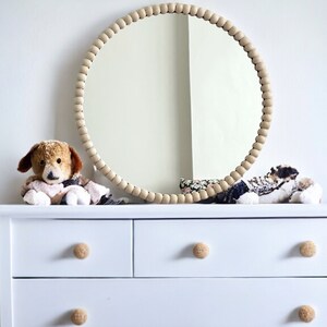 May include: A white dresser with a round mirror and a beaded wooden frame. The mirror is above the dresser, which has three drawers with round, light-colored knobs. Two stuffed animal dogs are on the dresser.