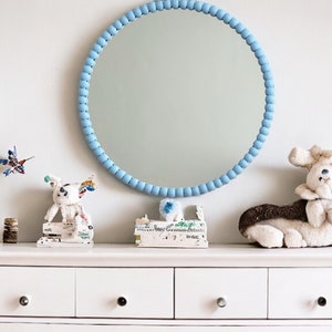 May include: A round mirror with a light blue beaded frame hangs above a white dresser. The dresser is decorated with a variety of small objects, including a stuffed animal and a small sculpture. The wall is painted a light gray color.