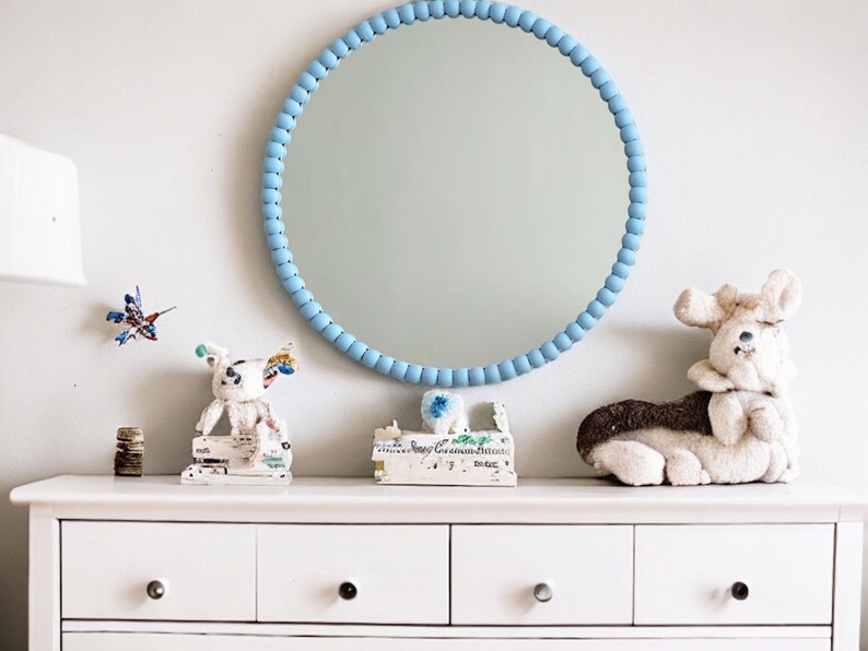 May include: A round mirror with a light blue beaded frame hangs above a white dresser. The dresser is decorated with a variety of small objects, including a stuffed animal and a small sculpture. The wall is painted a light gray color.