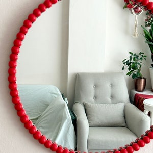 May include: A round mirror with a red beaded frame. The mirror reflects a light gray armchair with a pillow, a sofa, and plants. The mirror is mounted on a white wall, adding a decorative touch to the room.