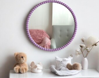 Op de afbeelding: Een ronde spiegel met een paarsgekleurde kralenlijst. De spiegel hangt aan een witte muur boven een witte commode. Op de commode staan een teddybeer, een wit keramiek voorwerp en een grijze gebreide muts.