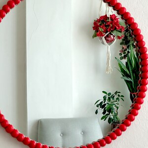 May include: A round mirror with a red beaded frame. The mirror reflects a white wall, a gray chair, and hanging plants with red flowers. The mirror is a decorative wall hanging.