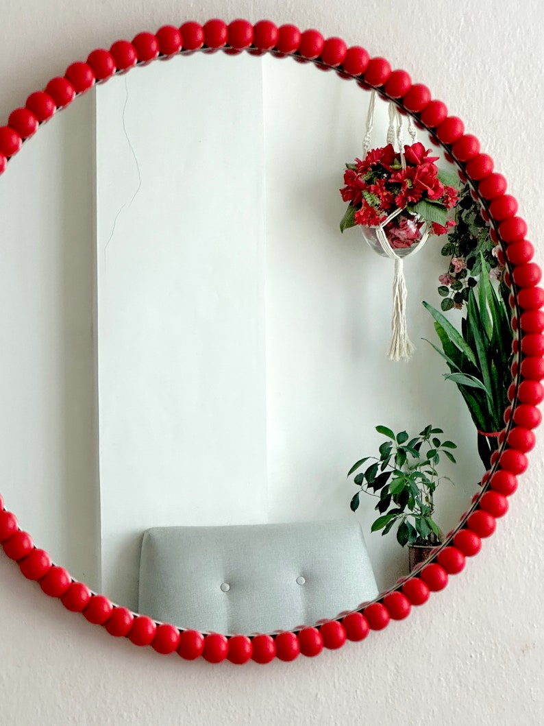 May include: A round mirror with a red beaded frame. The mirror reflects a white wall, a gray chair, and hanging plants with red flowers. The mirror is a decorative wall hanging.