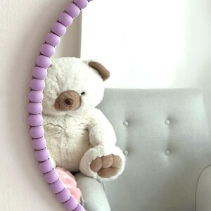May include: A round mirror with a lavender colored beaded frame. The mirror is reflecting a white teddy bear sitting in a light gray chair.