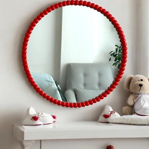May include: A round mirror with a red beaded frame hangs on a white wall. The mirror reflects a gray armchair and a window. A white dresser holds a teddy bear and two pairs of white baby shoes with red accents.