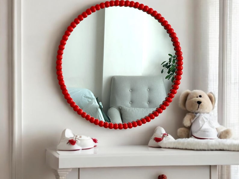 May include: A round mirror with a red beaded frame hangs on a white wall. The mirror reflects a gray armchair and a window. A white dresser holds a teddy bear and two pairs of white baby shoes with red accents.