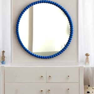 May include: A round mirror with a vibrant blue beaded frame hangs above a white dresser. The mirror reflects the room, and the dresser has gold-colored knobs. Decorative items sit on the dresser.