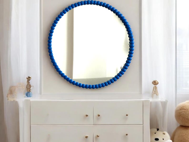 May include: A round mirror with a vibrant blue beaded frame hangs above a white dresser. The mirror reflects the room, and the dresser has gold-colored knobs. Decorative items sit on the dresser.