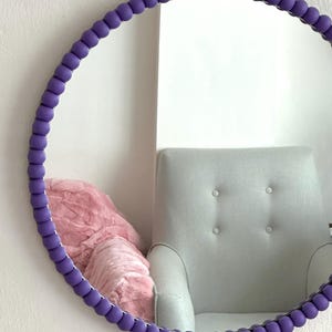 May include: A round mirror with a purple beaded frame. The mirror is reflecting a chair with a pink throw blanket.